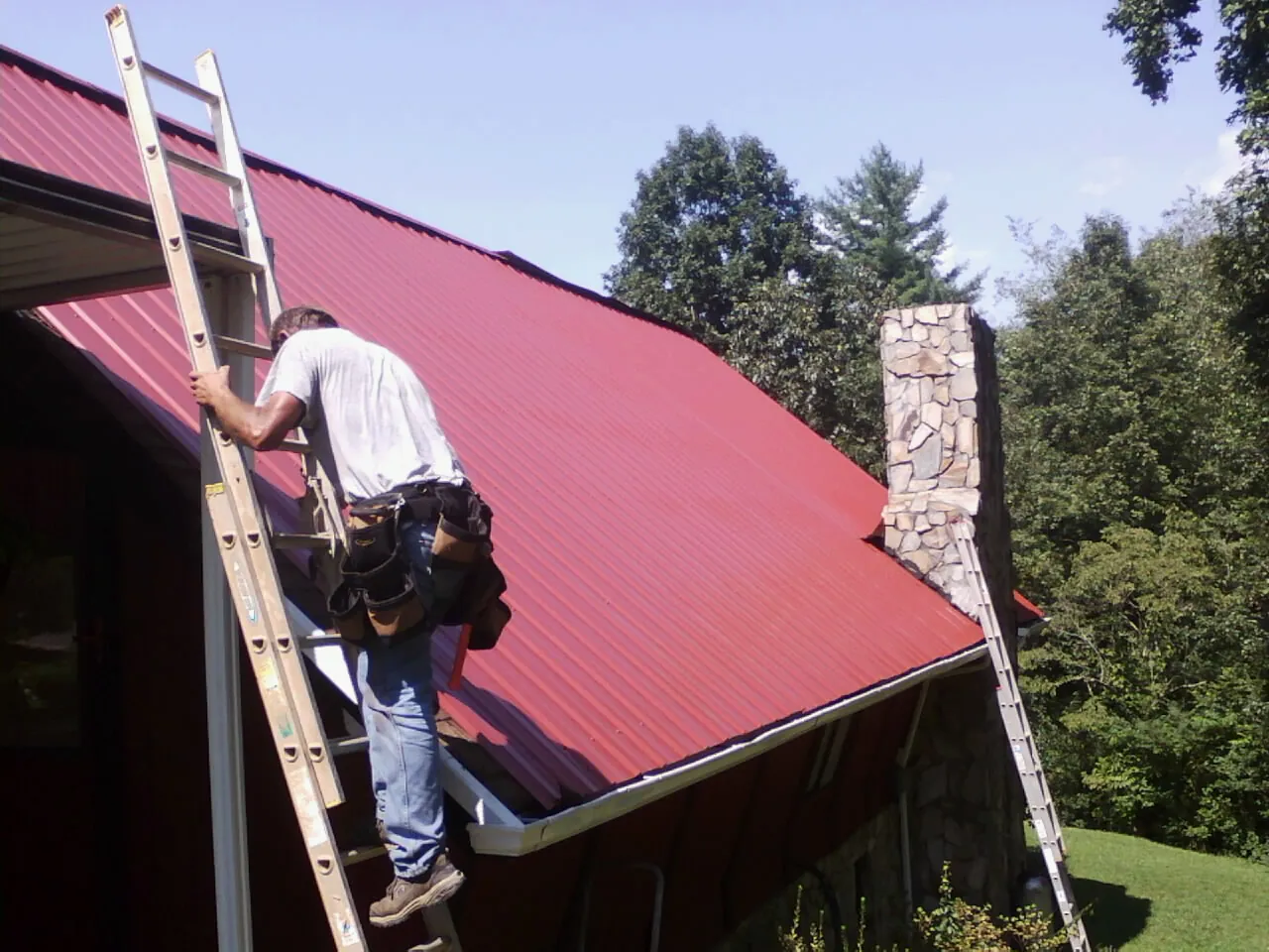 Roofer on a ladder beside a red metal roof and stone chimney — Soffit & Fascia Repair by Roofing Brothers in Pulteney